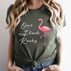 K&ouml;nnte beinhalten: Olivgr&uuml;nes T-Shirt mit dem wei&szlig;en Schriftzug "Our Flock Rocks". Auf der rechten Seite befindet sich eine rosa Flamingo-Grafik. Das Shirt hat einen Rundhalsausschnitt.