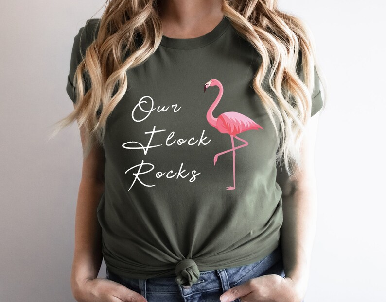 K&ouml;nnte beinhalten: Olivgr&uuml;nes T-Shirt mit dem wei&szlig;en Schriftzug "Our Flock Rocks". Auf der rechten Seite befindet sich eine rosa Flamingo-Grafik. Das Shirt hat einen Rundhalsausschnitt.