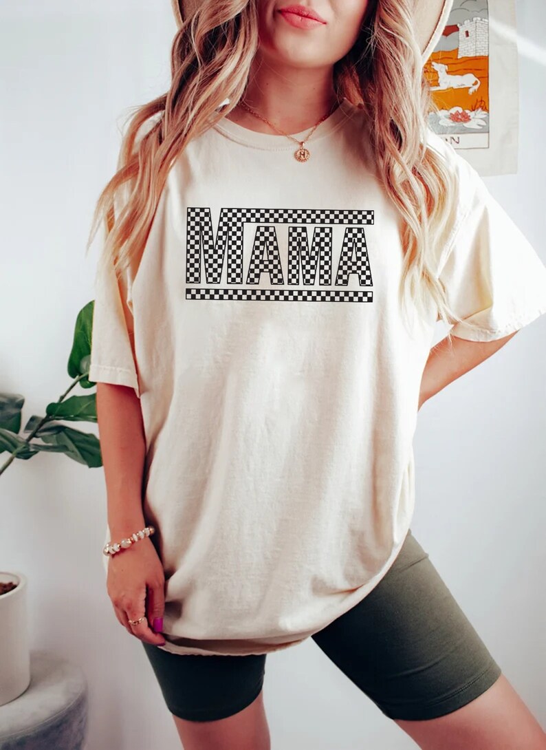 Puede incluir: Camiseta color crema con la palabra "MAMA" en un dise&ntilde;o a cuadros blanco y negro. La camiseta es de corte holgado y tiene mangas cortas. La modelo lleva un pantal&oacute;n corto verde oscuro y una pulsera.