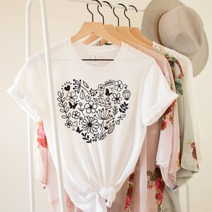 May include: White t-shirt with a black floral heart design. The heart is made up of flowers, leaves, and butterflies.