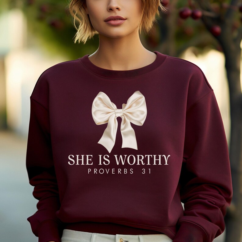 May include: A maroon sweatshirt with a large cream-colored bow graphic. The text "SHE IS WORTHY PROVERBS 31" is printed below the bow in white. The sweatshirt is a solid color and has a crew neck.