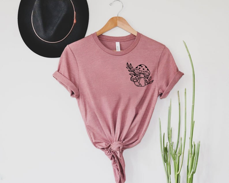 Peut inclure: T-shirt de couleur rose poudr&eacute; avec un motif de champignon noir sur le devant. Le motif repr&eacute;sente deux champignons et des feuilles. Le t-shirt est suspendu &agrave; un cintre en bois sur fond blanc.