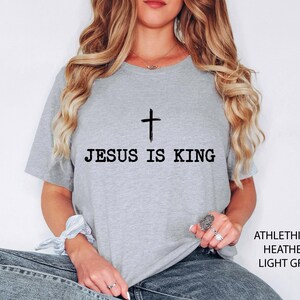 K&ouml;nnte beinhalten: Hellgraues T-Shirt mit dem schwarzen Text "JESUS IS KING", dar&uuml;ber ein schwarzes Kreuz. Das T-Shirt besteht aus Athlethich Heather Light Grey Material. Das T-Shirt hat einen Rundhalsausschnitt.
