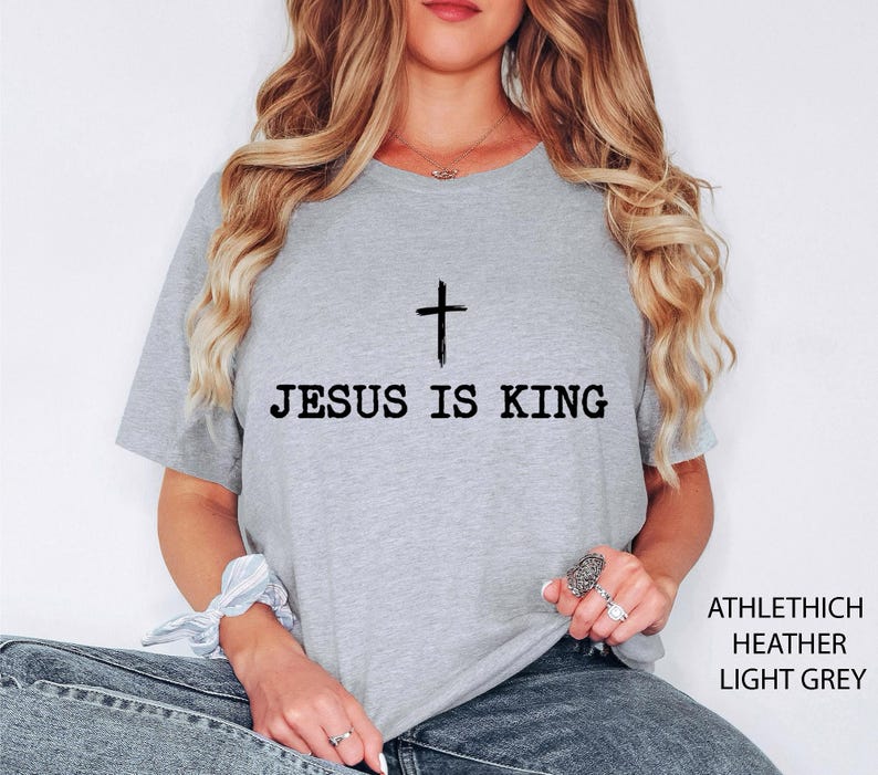 K&ouml;nnte beinhalten: Hellgraues T-Shirt mit dem schwarzen Text "JESUS IS KING", dar&uuml;ber ein schwarzes Kreuz. Das T-Shirt besteht aus Athlethich Heather Light Grey Material. Das T-Shirt hat einen Rundhalsausschnitt.