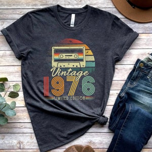 May include: A dark heather grey short-sleeve t-shirt featuring a graphic of a cassette tape centred above a sunset design. The text reads "Vintage 1976 LIMITED EDITION" in a retro font. The graphic uses orange, yellow, and teal colour stripes.