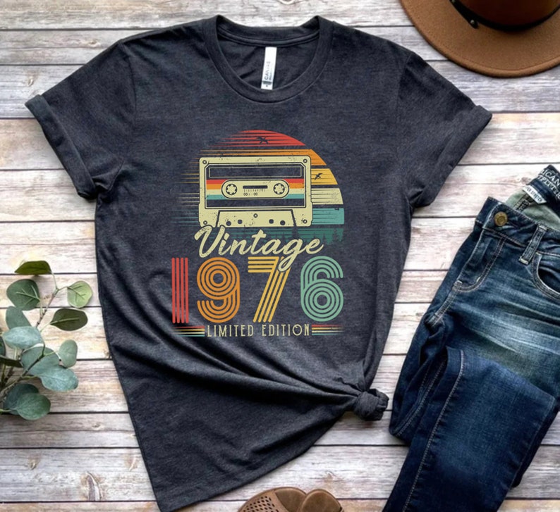 May include: A dark heather grey short-sleeve t-shirt featuring a graphic of a cassette tape centred above a sunset design. The text reads "Vintage 1976 LIMITED EDITION" in a retro font. The graphic uses orange, yellow, and teal colour stripes.