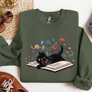 May include: A green sweatshirt with a black cat reading a book surrounded by flowers. The cat is in a field of flowers with butterflies and a hummingbird.