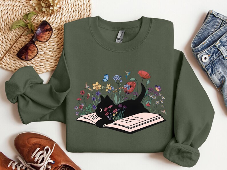 May include: A green sweatshirt with a black cat reading a book surrounded by flowers. The cat is in a field of flowers with butterflies and a hummingbird.
