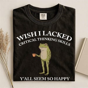 May include: A black crew neck t-shirt featuring a graphic of a green frog holding a small brown mug. The text above the frog reads WISH I LACKED CRITICAL THINKING SKILLS and the text below the frog reads Y'ALL SEEM SO HAPPY in white lettering.