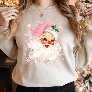 May include: A beige sweatshirt featuring a vintage Santa Claus graphic with a pink hat and snowflakes. The Santa has a rosy face and white beard. The design is a retro Christmas theme.