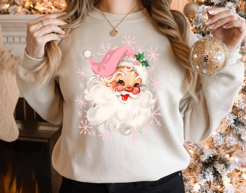 May include: A beige sweatshirt featuring a vintage Santa Claus graphic with a pink hat and snowflakes. The Santa has a rosy face and white beard. The design is a retro Christmas theme.