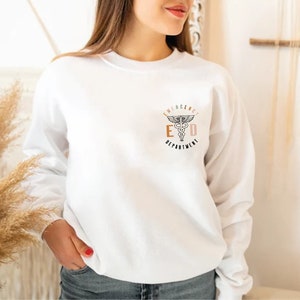 May include: White sweatshirt with a small graphic on the upper left chest. The graphic features a medical symbol with the text "EMERGENCY DEPARTMENT" in a stylized font. The sweatshirt is long-sleeved and appears to be made of a soft material.