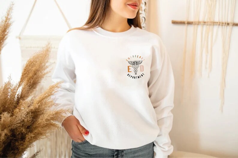 May include: White sweatshirt with a small graphic on the upper left chest. The graphic features a medical symbol with the text "EMERGENCY DEPARTMENT" in a stylized font. The sweatshirt is long-sleeved and appears to be made of a soft material.