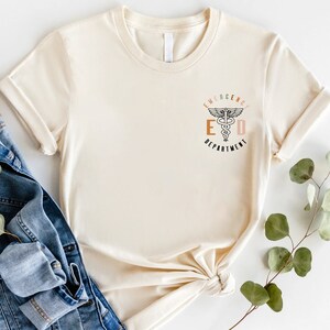 May include: Cream-colored t-shirt with rolled sleeves. The shirt features the text "EMERGENCY DEPARTMENT" with a medical symbol in the center. The text is in a mix of colors, including orange, brown, and pink. The shirt is paired with a denim jacket and greenery.