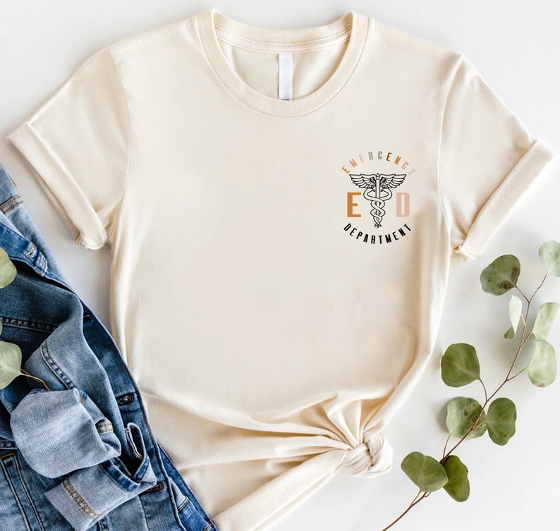 May include: Cream-colored t-shirt with rolled sleeves. The shirt features the text "EMERGENCY DEPARTMENT" with a medical symbol in the center. The text is in a mix of colors, including orange, brown, and pink. The shirt is paired with a denim jacket and greenery.