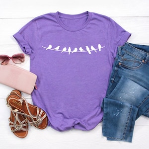 May include: Purple t-shirt with a white graphic of birds on a clothesline. The shirt is laid out on a white surface with a pair of pink sunglasses, a pink wallet, and a pair of brown sandals.