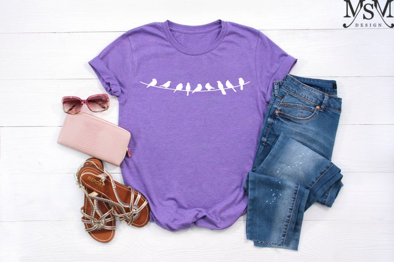 May include: Purple t-shirt with a white graphic of birds on a clothesline. The shirt is laid out on a white surface with a pair of pink sunglasses, a pink wallet, and a pair of brown sandals.