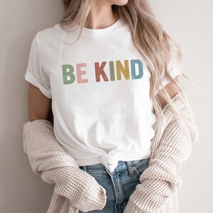 May include: White t-shirt with the words "BE KIND" in colorful, block letters. The shirt is paired with blue jeans and a cream-colored sweater. The text is in green, pink, yellow, and blue.