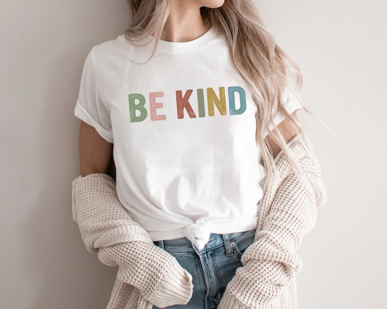 May include: White t-shirt with the words "BE KIND" in colorful, block letters. The shirt is paired with blue jeans and a cream-colored sweater. The text is in green, pink, yellow, and blue.