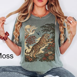 May include: A moss green short-sleeve t-shirt featuring a graphic print of a tiger leaping among flowers, trees, clouds, a bird, and a butterfly. The text Moss is printed on the left side. The shirt has a crew neckline and a relaxed fit design.