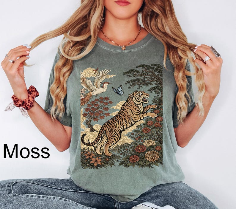 May include: A moss green short-sleeve t-shirt featuring a graphic print of a tiger leaping among flowers, trees, clouds, a bird, and a butterfly. The text Moss is printed on the left side. The shirt has a crew neckline and a relaxed fit design.