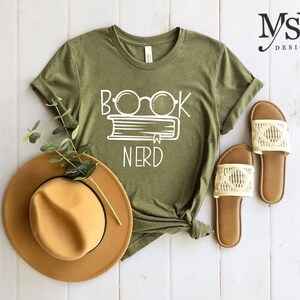 May include: Olive green t-shirt with the white text "BOOK NERD" above a book and glasses graphic. A tan fedora hat and a pair of cream-colored sandals are next to the shirt. The shirt is knotted at the bottom.