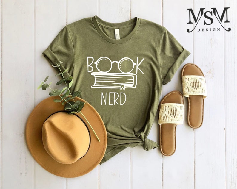 May include: Olive green t-shirt with the white text "BOOK NERD" above a book and glasses graphic. A tan fedora hat and a pair of cream-colored sandals are next to the shirt. The shirt is knotted at the bottom.
