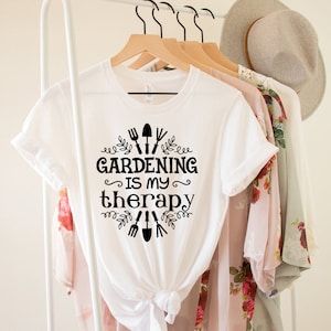 Op de afbeelding: Wit T-shirt met zwarte tekst die "Tuinieren is mijn therapie" zegt met een afbeelding van tuingereedschap en bladeren.