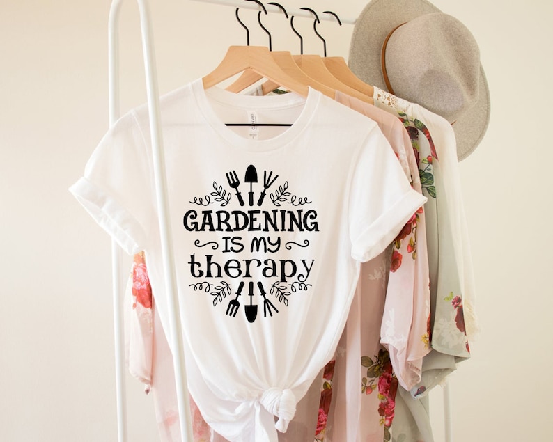 Op de afbeelding: Wit T-shirt met zwarte tekst die "Tuinieren is mijn therapie" zegt met een afbeelding van tuingereedschap en bladeren.