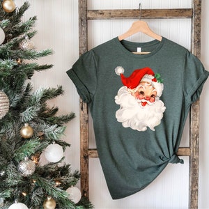 May include: A dark green t-shirt featuring a vintage Santa Claus graphic. The Santa Claus illustration has a red hat, white beard, and rosy cheeks. The shirt is hanging on a wooden hanger against a rustic ladder.