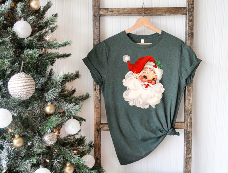 May include: A dark green t-shirt featuring a vintage Santa Claus graphic. The Santa Claus illustration has a red hat, white beard, and rosy cheeks. The shirt is hanging on a wooden hanger against a rustic ladder.