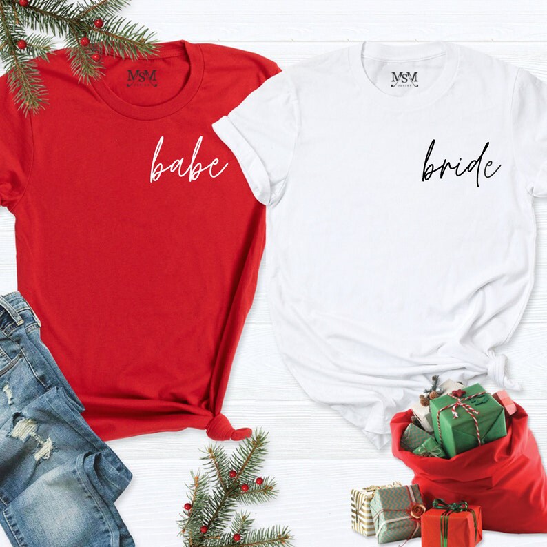 K&ouml;nnte beinhalten: Zwei kurz&auml;rmelige T-Shirts, eines rot und eines wei&szlig;. Das rote Shirt hat das Wort "babe" in Schreibschrift, und das wei&szlig;e Shirt hat das Wort "bride" in Schreibschrift. Die Shirts liegen auf einer wei&szlig;en Oberfl&auml;che mit Weihnachtsdekorationen.