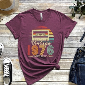 May include: A maroon short-sleeve t-shirt featuring a graphic of a cassette tape centred above the text "Vintage 1976 LIMITED EDITION". The design uses a retro colour palette of orange, yellow, and teal stripes set against a circular background.