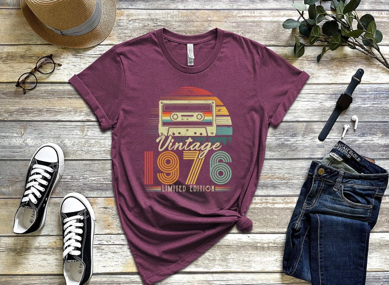 May include: A maroon short-sleeve t-shirt featuring a graphic of a cassette tape centred above the text "Vintage 1976 LIMITED EDITION". The design uses a retro colour palette of orange, yellow, and teal stripes set against a circular background.