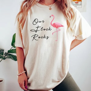 K&ouml;nnte beinhalten: Cremefarbenes T-Shirt mit dem Text "Our Flock Rocks" und einer rosa Flamingo-Grafik. Das Shirt hat eine lockere Passform und kurze &Auml;rmel. Der Flamingo steht auf einem Bein.