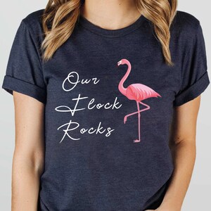 K&ouml;nnte beinhalten: Dunkelmarineblaues T-Shirt mit einer rosa Flamingo-Grafik und dem Text "Our Flock Rocks" in wei&szlig;er Schrift. Der Flamingo steht auf einem Bein. Das T-Shirt hat hochgekrempelte &Auml;rmel.
