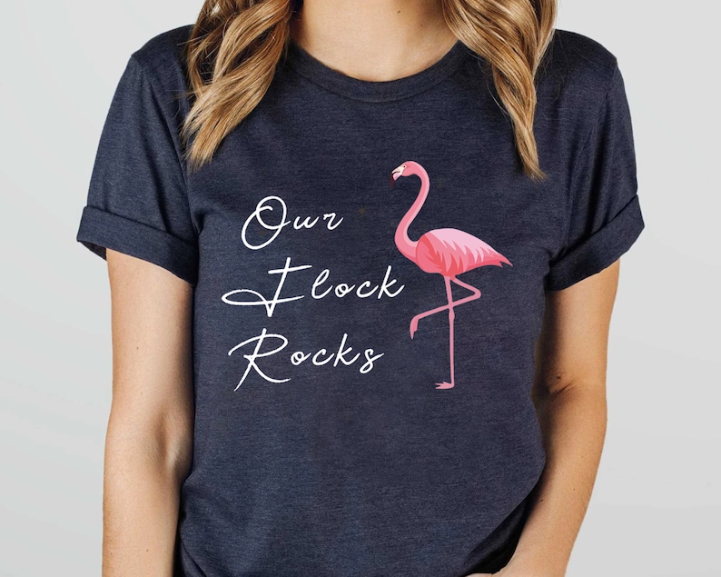 K&ouml;nnte beinhalten: Dunkelmarineblaues T-Shirt mit einer rosa Flamingo-Grafik und dem Text "Our Flock Rocks" in wei&szlig;er Schrift. Der Flamingo steht auf einem Bein. Das T-Shirt hat hochgekrempelte &Auml;rmel.