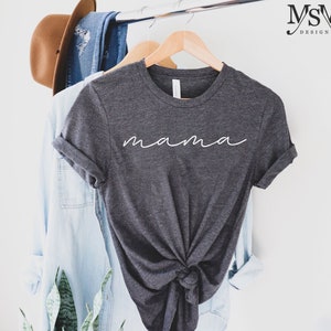 Puede incluir: Camiseta gris oscuro con la palabra "mama" en escritura blanca. La camiseta est&aacute; anudada en la parte inferior y cuelga de una percha de madera. Una camisa vaquera azul claro y un sombrero marr&oacute;n son visibles en el fondo.