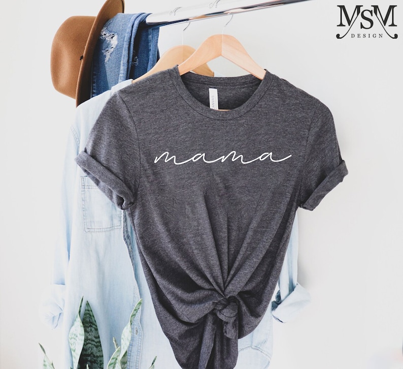 Puede incluir: Camiseta gris oscuro con la palabra "mama" en escritura blanca. La camiseta est&aacute; anudada en la parte inferior y cuelga de una percha de madera. Una camisa vaquera azul claro y un sombrero marr&oacute;n son visibles en el fondo.