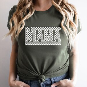 Puede incluir: Una camiseta verde oliva con la palabra "MAMA" en un patr&oacute;n blanco a cuadros. El texto est&aacute; centrado en la camiseta, con el dise&ntilde;o a cuadros que tambi&eacute;n delinea la palabra. Una prenda casual para el d&iacute;a a d&iacute;a.