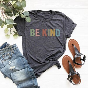 May include: Dark gray t-shirt with the words "BE KIND" in colorful letters. The shirt is paired with blue jeans and brown sandals. A sprig of greenery is in the background.
