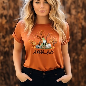 May include: A burnt orange t-shirt featuring a graphic print of a gnome, a black cat, and pumpkins nestled among autumn trees. The text "Ahhhh...fall" is printed in black script below the illustration. The shirt has short sleeves and a crew neckline.
