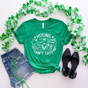 May include: Green t-shirt with a white graphic that says "Hoeing Ain't Easy" and features gardening tools and plants. The shirt is paired with denim shorts and black shoes. Green garland is around the edges of the image.