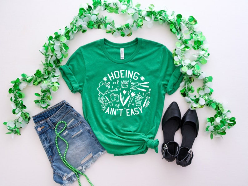 May include: Green t-shirt with a white graphic that says "Hoeing Ain't Easy" and features gardening tools and plants. The shirt is paired with denim shorts and black shoes. Green garland is around the edges of the image.
