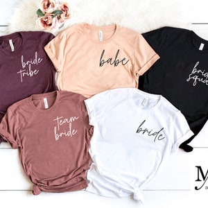 May include: Five different t-shirts with various bridal party slogans. The shirts are in different colors, including maroon, peach, white, black, and brown. The slogans on the shirts include "bride tribe", "babe", "bride squad", "team bride", and "bride".