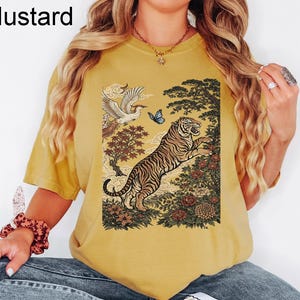 May include: A mustard yellow short-sleeved t-shirt featuring a graphic print of a tiger leaping among flowers and trees, with a bird and a blue butterfly above. The text "Mustard" is printed in the upper left corner of the image.