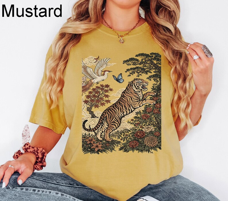 May include: A mustard yellow short-sleeved t-shirt featuring a graphic print of a tiger leaping among flowers and trees, with a bird and a blue butterfly above. The text "Mustard" is printed in the upper left corner of the image.