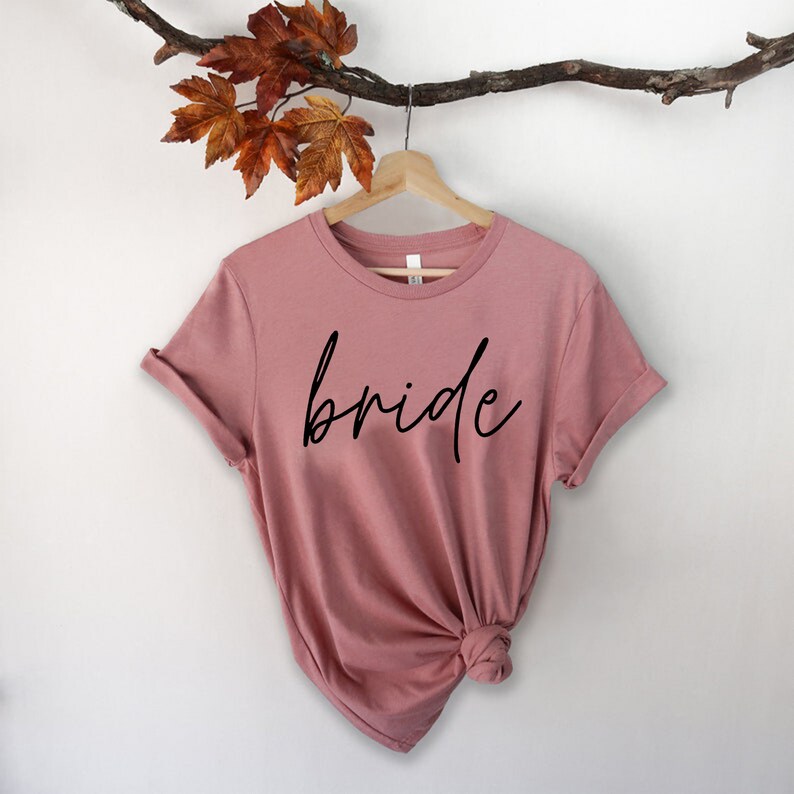 May include: A dusty rose-colored t-shirt with the word "bride" in black script. The shirt is hanging on a wooden hanger, with a branch and leaves in the background. The t-shirt has rolled-up sleeves.