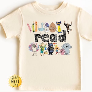 May include: A cream-colored short-sleeve t-shirt featuring the word read in a black scribbled font. Above and below the text are colorful illustrations of various children's book characters, including crayons, a pigeon, a monkey, and an elephant.