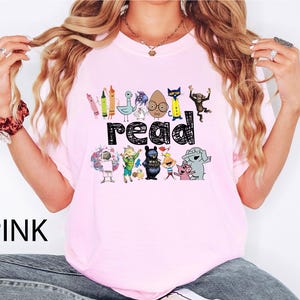 May include: A light pink t-shirt featuring the word read in a black scribbled font. The design includes colorful illustrations of various children's book characters, including a pigeon, a cat, a monkey, an elephant, and a pig, arranged above and below the text.